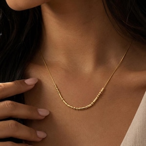 Puede incluir: Un collar de tono dorado que presenta una cadena delicada con un segmento central compuesto por pequeñas cuentas cilíndricas y diminutas cuentas esféricas dispuestas en un patrón que recuerda al código Morse, sobre la clavícula.