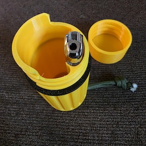 May include: A yellow 3D-printed lighter case with a black band and a green cord. The case holds a silver lighter. The lid is off to the side. The case is on a dark brown textured surface.