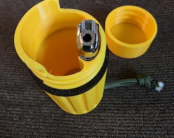 May include: A yellow 3D-printed lighter case with a black band and a green cord. The case holds a silver lighter. The lid is off to the side. The case is on a dark brown textured surface.
