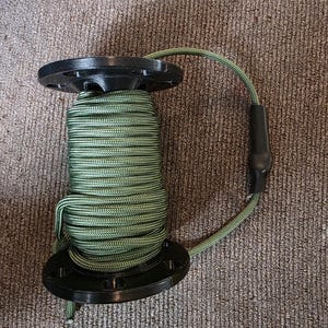 May include: A spool of green and black cord wrapped around a black plastic reel. The reel has a circular design with multiple holes. A length of cord extends from the spool, connected to a black plastic piece.