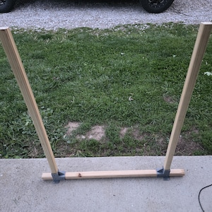 May include: A wooden kayak or canoe stand. The stand is made of light-colored wood and metal brackets. The stand has two angled uprights connected by a horizontal base. The stand is on a concrete surface with grass in the background.