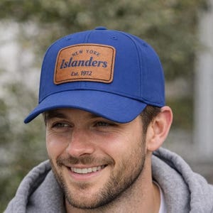 Op de afbeelding: Een koningsblauwe baseballcap met een gebogen klep. Het heeft een bruin leren patch met de tekst "New York Islanders" en "Est. 1972" in marineblauwe letters. Een sportief accessoire.