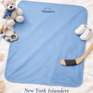 May include: A light blue infant blanket with the text "New York Islanders" in navy blue. The blanket is surrounded by a white teddy bear, a wooden hockey stick, a baby rattle, and a pair of white baby shoes.
