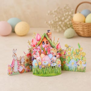 May include: A three-paneled decorative paper display featuring a central cottage surrounded by pink tulips, green grass, and colorful Easter eggs. Two rabbits are positioned near the house, while a third rabbit stands by a wooden fence on the left.