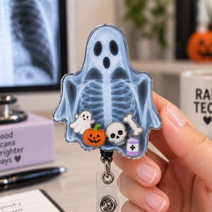 May include: A ghost-shaped badge reel featuring a blue and white X-ray skeleton print. The clear acrylic casing contains small floating charms including a white ghost, an orange pumpkin, a white skull, a white bone, and a purple medicine bottle.