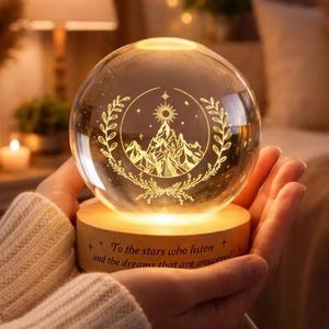 Pode incluir: Uma esfera de vidro transparente com um desenho gravado de montanhas e sol, repousando sobre uma base de madeira. A base tem o texto "To the stars who listen and the dreams that are answered!". A esfera é iluminada com um brilho quente.