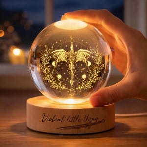 Pode incluir: Uma bola de cristal brilhante com um design de dragão dourado, repousando sobre uma base de madeira. A base é gravada com as palavras "Violent little thing" e uma espada. A esfera emite uma luz quente, criando um ambiente mágico.