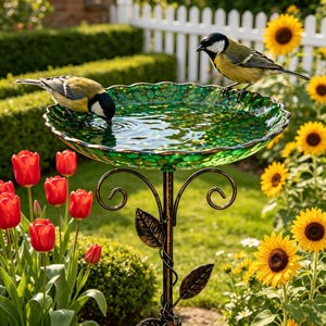 May include: A decorative green glass bird bath with a mosaic design, supported by a bronze-colored metal stand. Two small birds are perched on the edge of the bird bath. Red tulips and sunflowers are in the background.