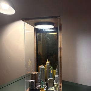 May include: A rectangular display case with a wooden frame and clear acrylic sides containing a miniature model of a city skyline. The display features various small buildings and a Statue of Liberty figure, illuminated by a circular light fixture.