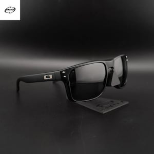 Pode incluir: Óculos de sol pretos com lentes escuras. Os óculos têm uma armação retangular e um pequeno logotipo prateado na haste. Os óculos são exibidos em um suporte preto.