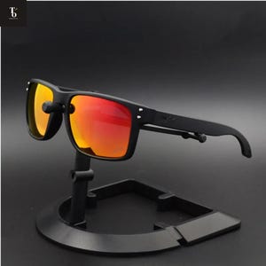 Pode incluir: Óculos de sol pretos com lentes laranja e vermelhas. Os óculos, de formato quadrado, são exibidos em um suporte preto. As lentes apresentam um efeito gradiente, passando do laranja para o vermelho.