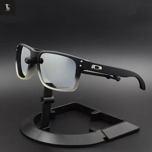 Puede incluir: Gafas de sol negras con lentes espejadas plateadas sobre un soporte negro. Las gafas, de montura cuadrada, presentan un degradado de negro a un color translúcido. Las lentes reflejan la luz.