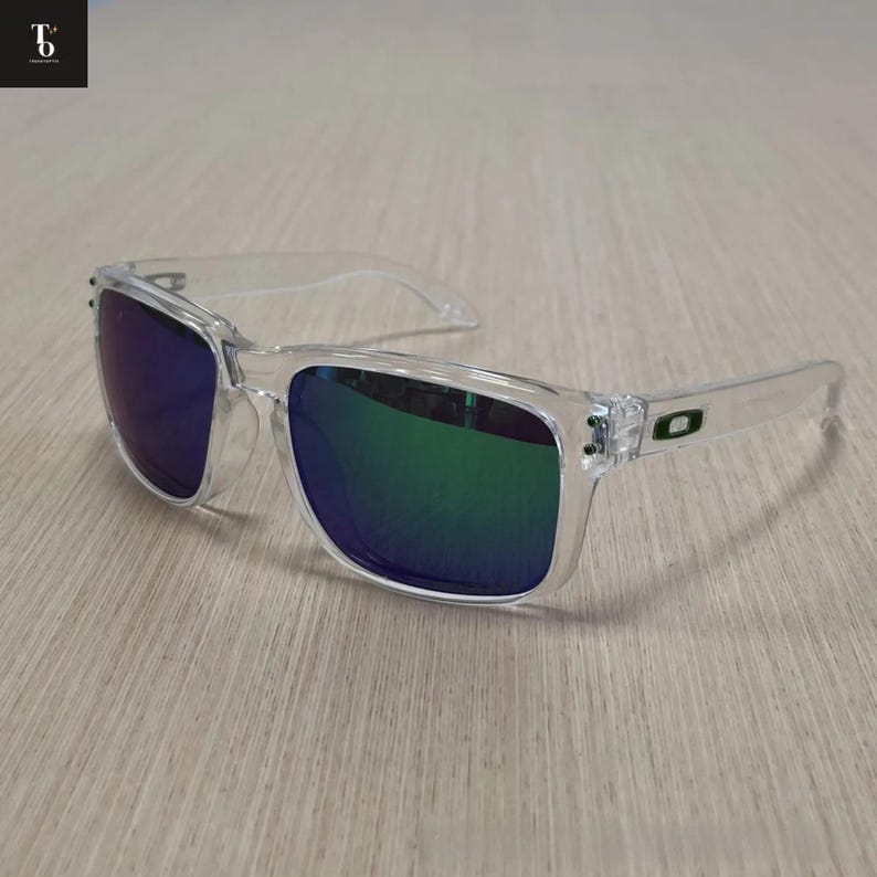 May include: Clear-framed sunglasses with mirrored lenses reflecting green and blue hues. The sunglasses have a rectangular shape and a subtle green logo on the temples. The frames are transparent, and the lenses are reflective.