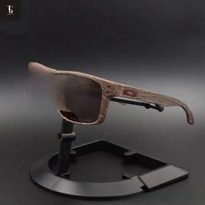 May include: Brown sunglasses with a wood grain pattern on the frames, displayed on a black stand. The lenses are a dark brown tint. The sunglasses have a rectangular shape and a small red logo on the side.