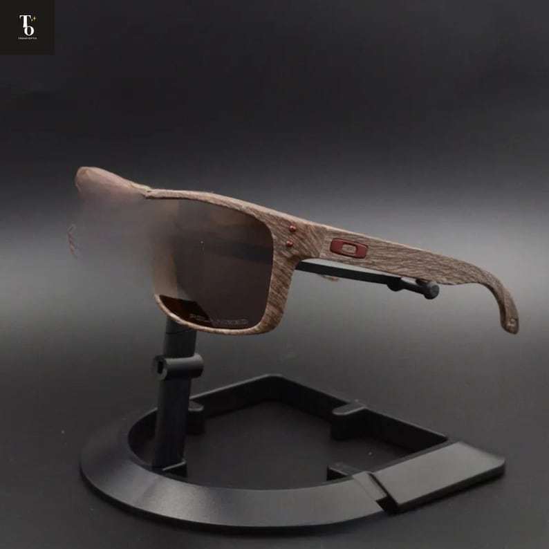 May include: Brown sunglasses with a wood grain pattern on the frames, displayed on a black stand. The lenses are a dark brown tint. The sunglasses have a rectangular shape and a small red logo on the side.