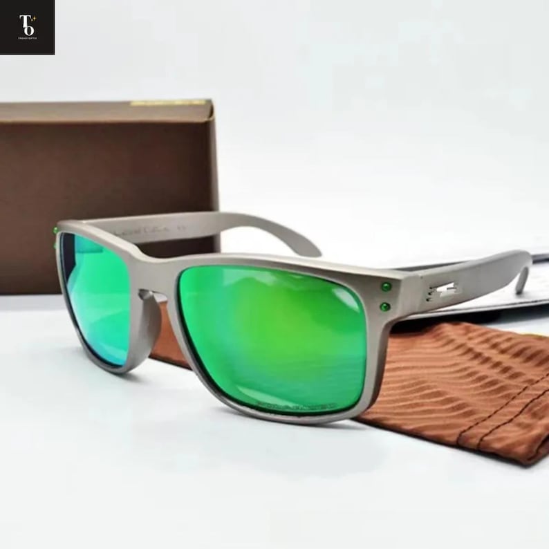 May include: Gray sunglasses with green lenses. The sunglasses have a rectangular frame and a brown carrying case. The lenses reflect a bright green color. A brown pouch is also included.