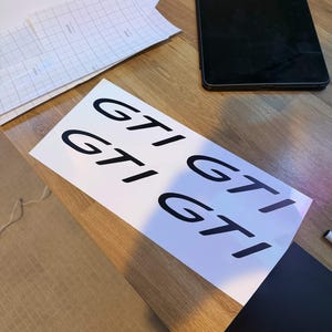 Op de afbeelding: Een wit vel papier met de zwarte tekst GTI vier keer in een raster gedrukt. In de buurt liggen vellen transferpapier met een raster en een zwarte tablet, allemaal gerangschikt op een houten oppervlak met een licht getextureerd gedeelte.