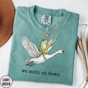 Puede incluir: Camiseta verde claro con un diseño caprichoso de una rana montada en un cisne, sosteniendo un lápiz. El ala del cisne presenta símbolos matemáticos. El texto "we math at dawn" está impreso debajo. También se ve un collar plateado con un colgante.