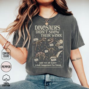 May include: A charcoal gray short-sleeve t-shirt featuring a graphic print with dinosaur skeletons and mathematical symbols. The text reads: DINOSAURS DIDN'T SHOW THEIR WORK and we all know what happened to them, along with labels like Forget the Negative.