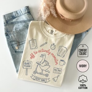 May include: Ivory-colored Comfort Colors t-shirt with a graphic of a goose on a scooter, surrounded by school-themed illustrations and text that reads "off to actively monitor" and "Test Day". The shirt is paired with light blue jeans and a tan hat.