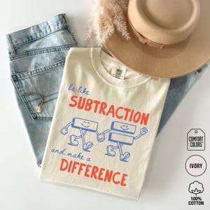May include: An ivory t-shirt featuring a graphic of a subtraction sign and an equals sign with arms and legs holding hands. The text reads, "be like SUBTRACTION and make a DIFFERENCE." The shirt is paired with light blue denim jeans and a tan hat.