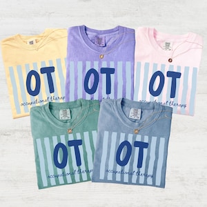 May include: Five short-sleeved t-shirts in yellow, purple, pink, green, and blue. Each shirt features a vertical light blue striped pattern, the bold dark blue letters OT, and the text occupational therapy printed in a cursive font below.
