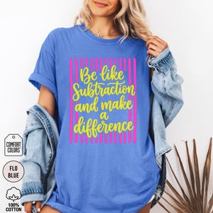 May include: A blue short-sleeved t-shirt featuring a graphic design with vertical pink stripes and yellow cursive text that reads: Be like Subtraction and make a difference. The shirt has a crew neckline and a relaxed fit, with a Comfort Colors label.