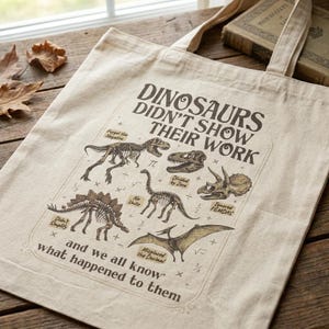 May include: A beige canvas tote bag featuring a dark brown graphic print of various dinosaur skeletons and fossils. The text reads: DINOSAURS DIDN'T SHOW THEIR WORK and we all know what happened to them, accompanied by small math symbols and labels.