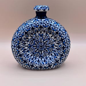 Op de afbeelding: Een decoratieve keramische fles met een ronde body en een kleine, cilindrische hals. De fles is wit met een gedetailleerd blauw bloemmotief. De hals en de basis zijn donkerblauw.