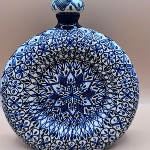 Può includere: Una bottiglia decorativa in ceramica con corpo arrotondato e tappo decorato. La bottiglia presenta un dettagliato design floreale blu e bianco, con un motivo centrale a stella. Il collo e il tappo sono di un blu più scuro.