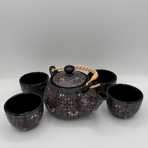 May include: A black teapot and four matching cups. The teapot has a decorative pattern in pink and gray, with a woven handle. The cups have a similar design. The set is designed for tea service.