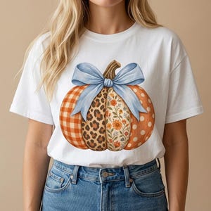 May include: A white short-sleeve t-shirt featuring a large graphic of a pumpkin. The pumpkin is divided into four vertical sections with orange gingham, leopard print, floral patterns, and orange polka dots, topped with a light blue bow.