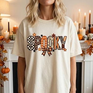 May include: A cream-colored short-sleeve t-shirt featuring the word SPOOKY printed in large, multi-patterned letters. The design includes black and white checkers, leopard print, orange plaid, and spiderweb motifs, accented by a plaid bow and stars.