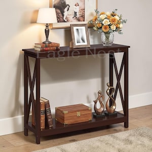 May include: A dark brown console table with an X-frame design. The table is adorned with a lamp, books, a framed photo, a vase of flowers, decorative sculptures, and a wooden box. The text "Eagle Nest" is visible on the table.