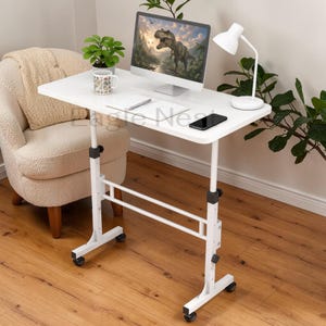 May include: A white adjustable laptop desk with a rectangular tabletop. The desk has a white frame with rolling wheels. A computer monitor, a white desk lamp, a smartphone, and a mug are on the desk. The background includes a beige armchair and a plant.
