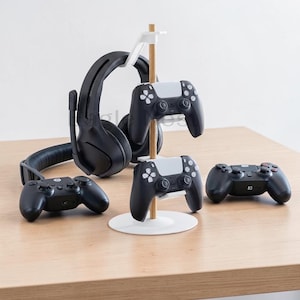 May include: A black gaming headset and four black game controllers are displayed on a light wood surface. Two controllers are mounted on a wooden and white stand. The controllers have white accents.