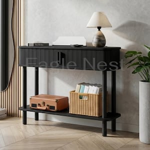 May include: A black console table with a fluted drawer front, a lower shelf, and four cylindrical legs. On top sits a table lamp with a beige shade and a stone-textured base. The lower shelf holds a tan rectangular case and a woven wicker basket.