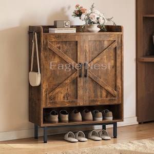 May include: A rustic brown wooden shoe cabinet with two doors and a lower shelf for shoes. The doors have a cross design. The cabinet has a shelf on top with a vase of flowers and a small radio. A purse hangs on the side.