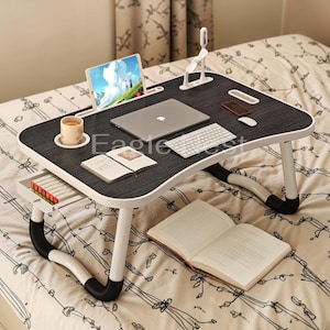 May include: A black and white laptop desk with a dark wood grain surface. The desk has a built-in tablet holder, a small drawer, and a cup holder. A laptop, tablet, keyboard, and other items are on the desk. The desk is on a bed.