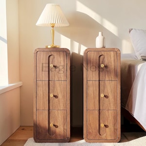 May include: Two tall, narrow bedside tables made of dark wood with rounded corners. Each table features three stacked drawers, each equipped with a small, circular gold-toned knob. The vertical design includes a recessed channel running down the center.