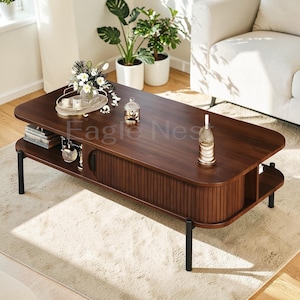 May include: A dark brown wooden coffee table with rounded edges, featuring a slatted tambour sliding door, an open storage shelf, and four thin black metal legs. The tabletop holds a decorative tray with flowers, a glass jar, and a bottle.