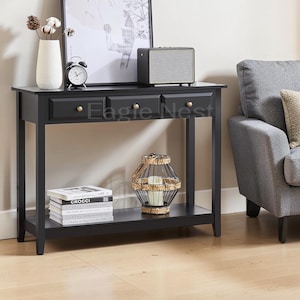 May include: A black rectangular console table with three drawers featuring small gold knobs. The table has a lower shelf holding a stack of books and a decorative wire lantern. The top surface holds a white vase, a clock, and a speaker.