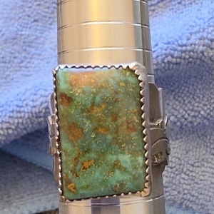 May include: A silver ring featuring a rectangular turquoise stone with brown matrix inclusions. The stone is set in a serrated bezel, and the silver band displays decorative stamped patterns on the sides, mounted on a metal ring mandrel.