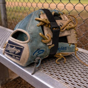 May include: A Rawlings baseball glove featuring a blue and tan leather exterior with tan lacing, a black adjustable strap across the top, and the number 19 written in blue ink on the side. The glove displays the Rawlings logo patch.