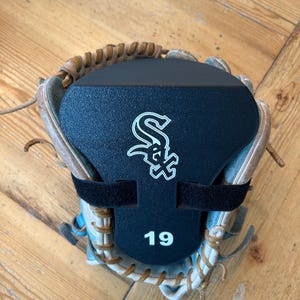 May include: A tan leather baseball glove featuring a black plastic form insert secured with a black strap. The insert displays the white Chicago White Sox logo and the number 19 in white text centered on the lower portion of the black surface.