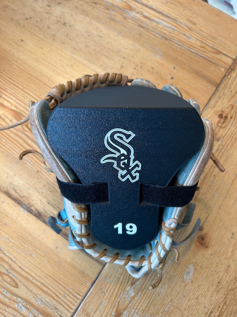 May include: A tan leather baseball glove featuring a black plastic form insert secured with a black strap. The insert displays the white Chicago White Sox logo and the number 19 in white text centered on the lower portion of the black surface.
