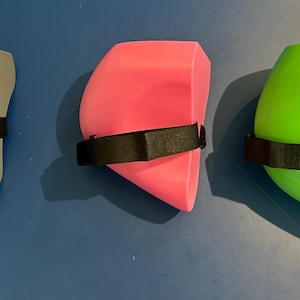 May include: Three protective knee pads in gray, bright pink, and lime green. Each pad features a smooth, curved, hard-shell exterior and a black adjustable fabric strap with a hook-and-loop fastener attached to the side for secure positioning.