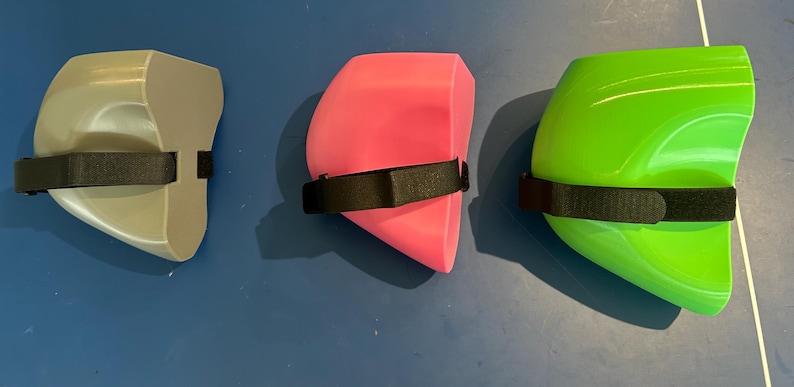 May include: Three protective knee pads in gray, bright pink, and lime green. Each pad features a smooth, curved, hard-shell exterior and a black adjustable fabric strap with a hook-and-loop fastener attached to the side for secure positioning.
