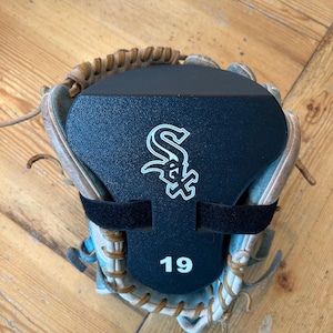 Peut inclure: Un gant de baseball en cuir beige avec un insert de forme en plastique noir fixé par une sangle noire. L'insert affiche le logo blanc des Chicago White Sox et le numéro 19 en texte blanc centré sur la partie inférieure de la surface noire.