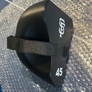 May include: A black foam knee pad featuring a white San Francisco Giants logo and the number 45 printed in white. The protective gear includes a black adjustable elastic strap with a hook and loop fastener for secure attachment during use.